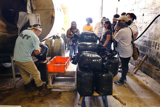 Picture of mushroom farm worker packing compost in black trash bags, surrounded by a group of onlookers, including Marcie, attentively watching the mushroom workers process
